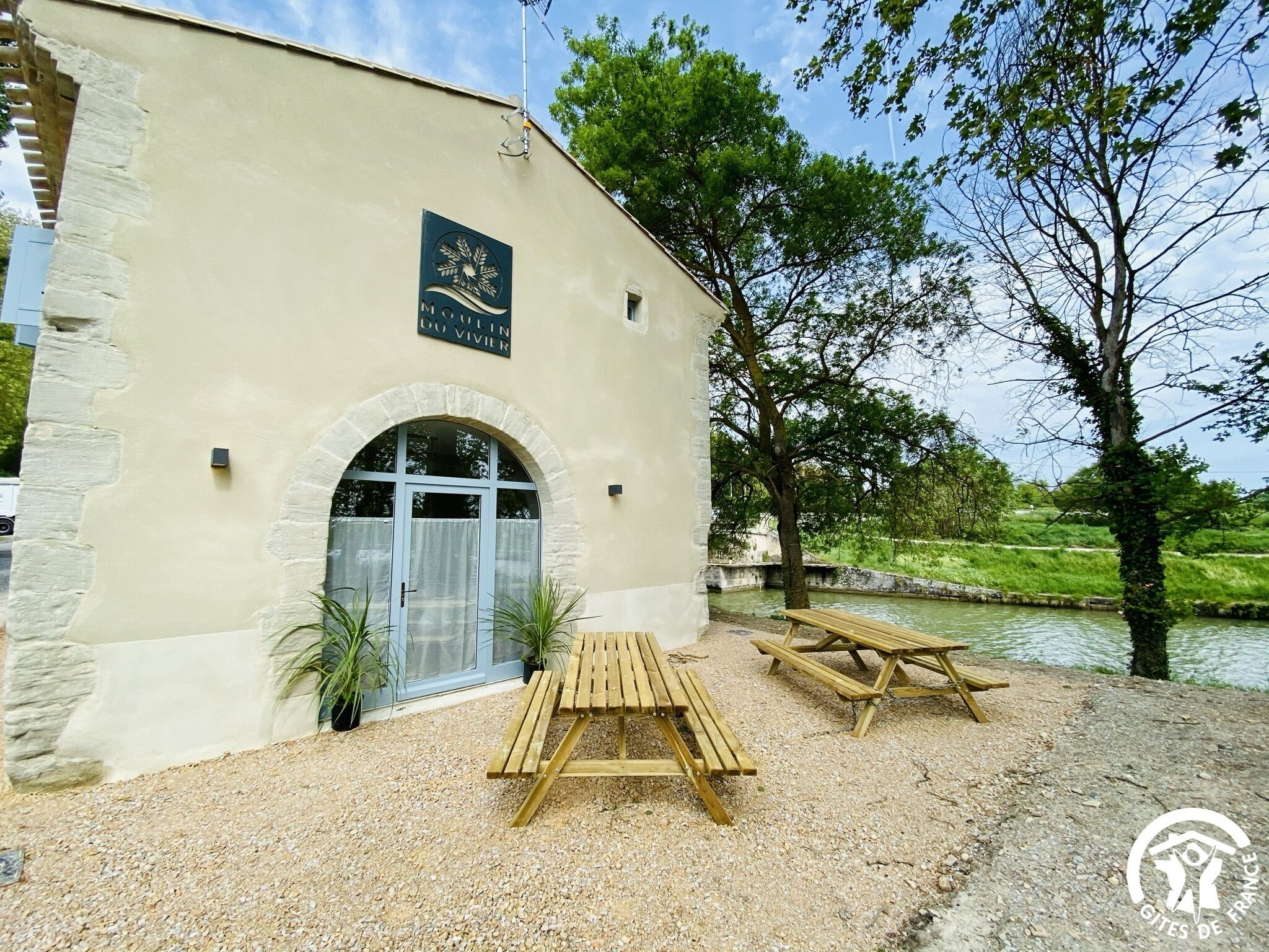 A building with a sign reading "Moulin du Vivier" and outdoor seating by a water body.