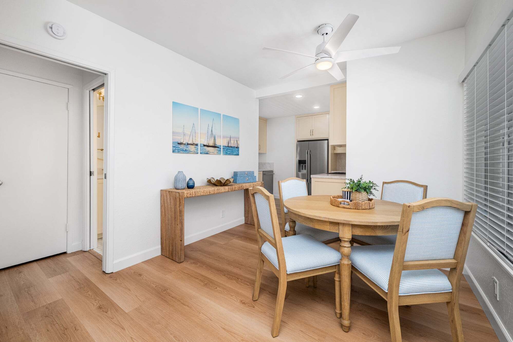 A dining area with a round wooden table, four chairs, a ceiling fan, and a kitchen in the background.