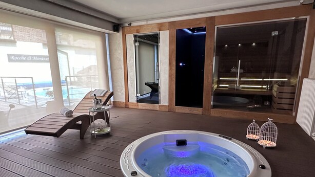 A hot tub on a wooden deck with a glass balustrade, a lounge chair, and a small table with a glass of water.
