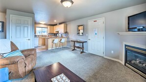 A living room with a brown leather couch, a wooden coffee table, and a fireplace. There is a kitchen in the background with wooden cabinets and a bar. - Apartment w/ Gas Fireplace ~ 1/2 Mi to Beach! (Anacortes)
