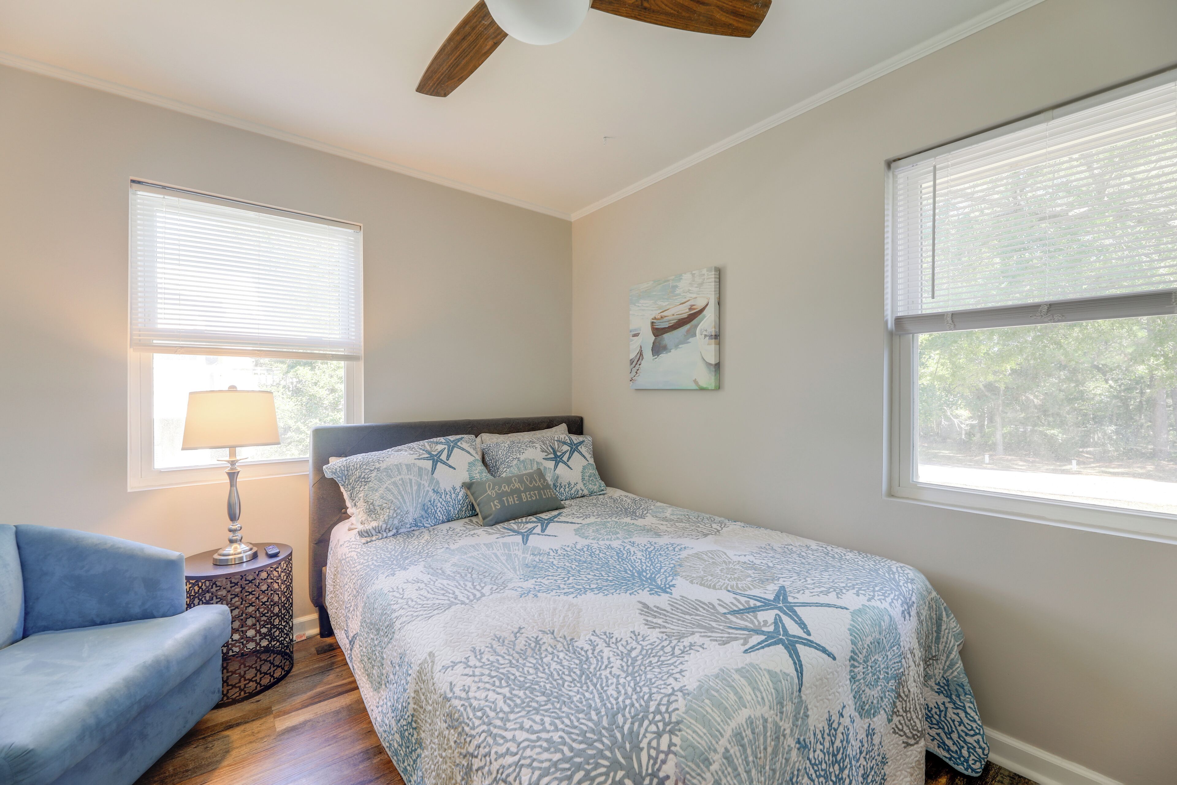 A bedroom with a bed, a nightstand with a lamp, a blue chair, and a ceiling fan.