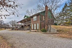 A two-story log cabin with a balcony, a gravel driveway, and a small garden area.