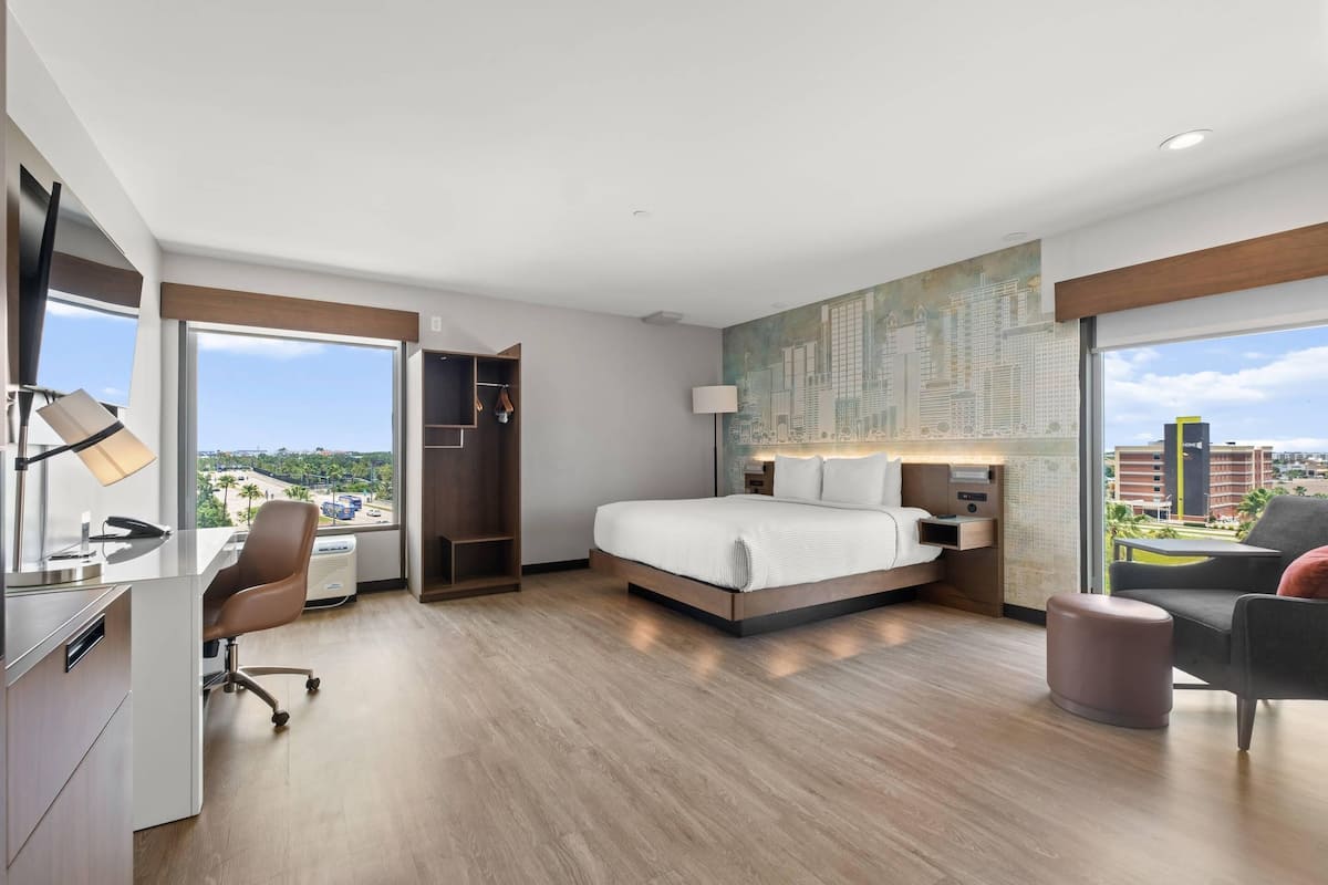 Habitación de hotel moderna con una cama grande, un escritorio y una silla. Se ve una ventana con vistas a la ciudad y un mural en la pared.