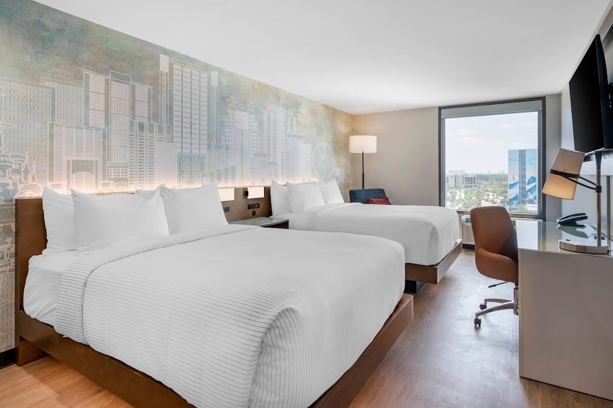 Habitación de hotel con dos camas, un escritorio, una silla, un ventanal con vistas a la ciudad y un mural en la pared.