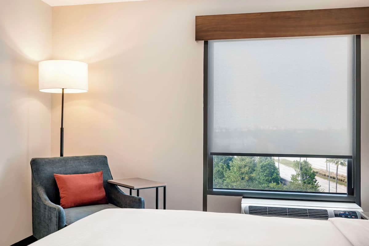 Habitación de hotel con cama, sillón con cojín, una mesita, lámpara y un ventanal con vistas a árboles y una carretera.