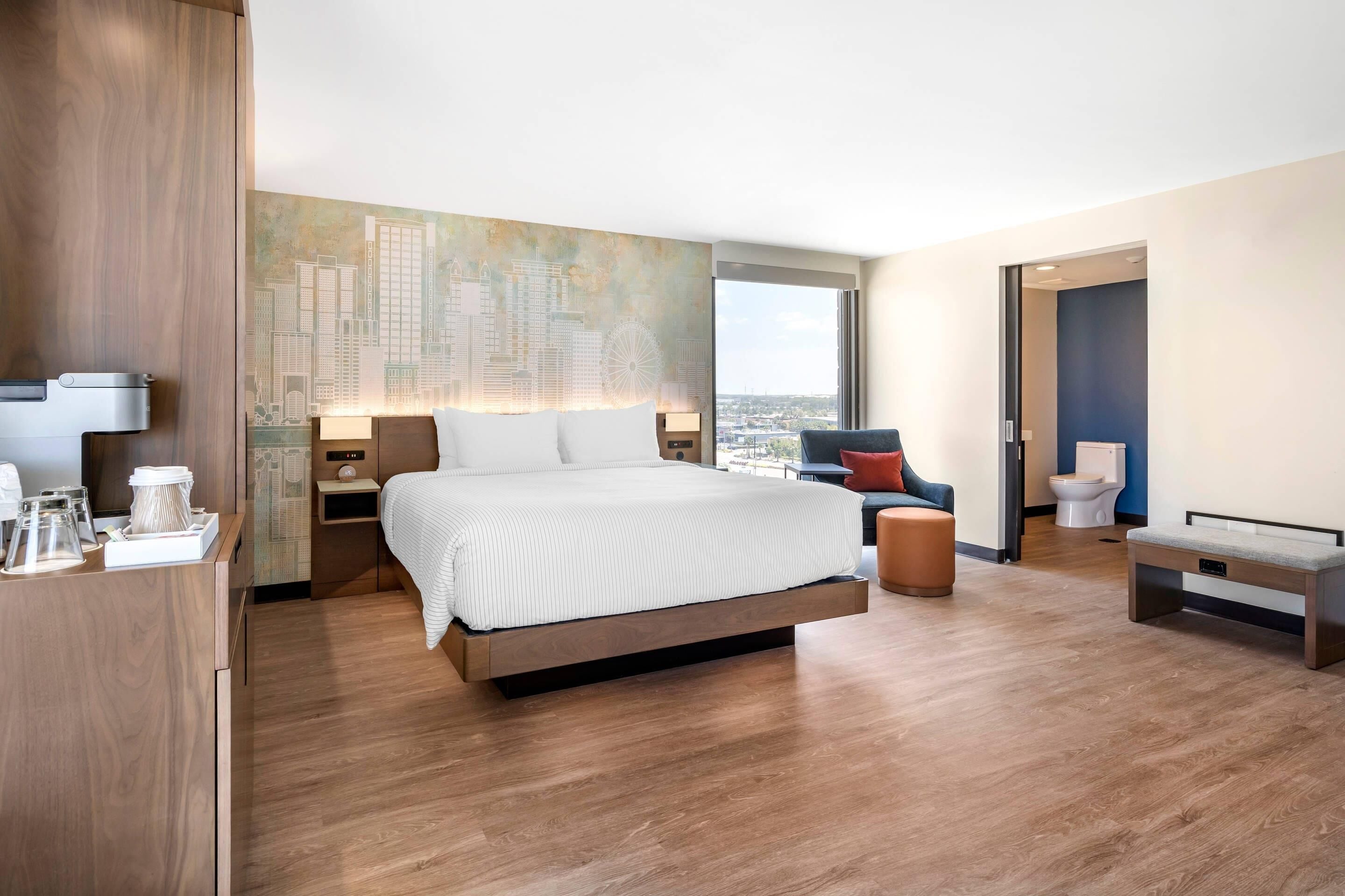 A modern hotel room with a large bed, a cityscape mural, a desk with a lamp, and a view of the city.