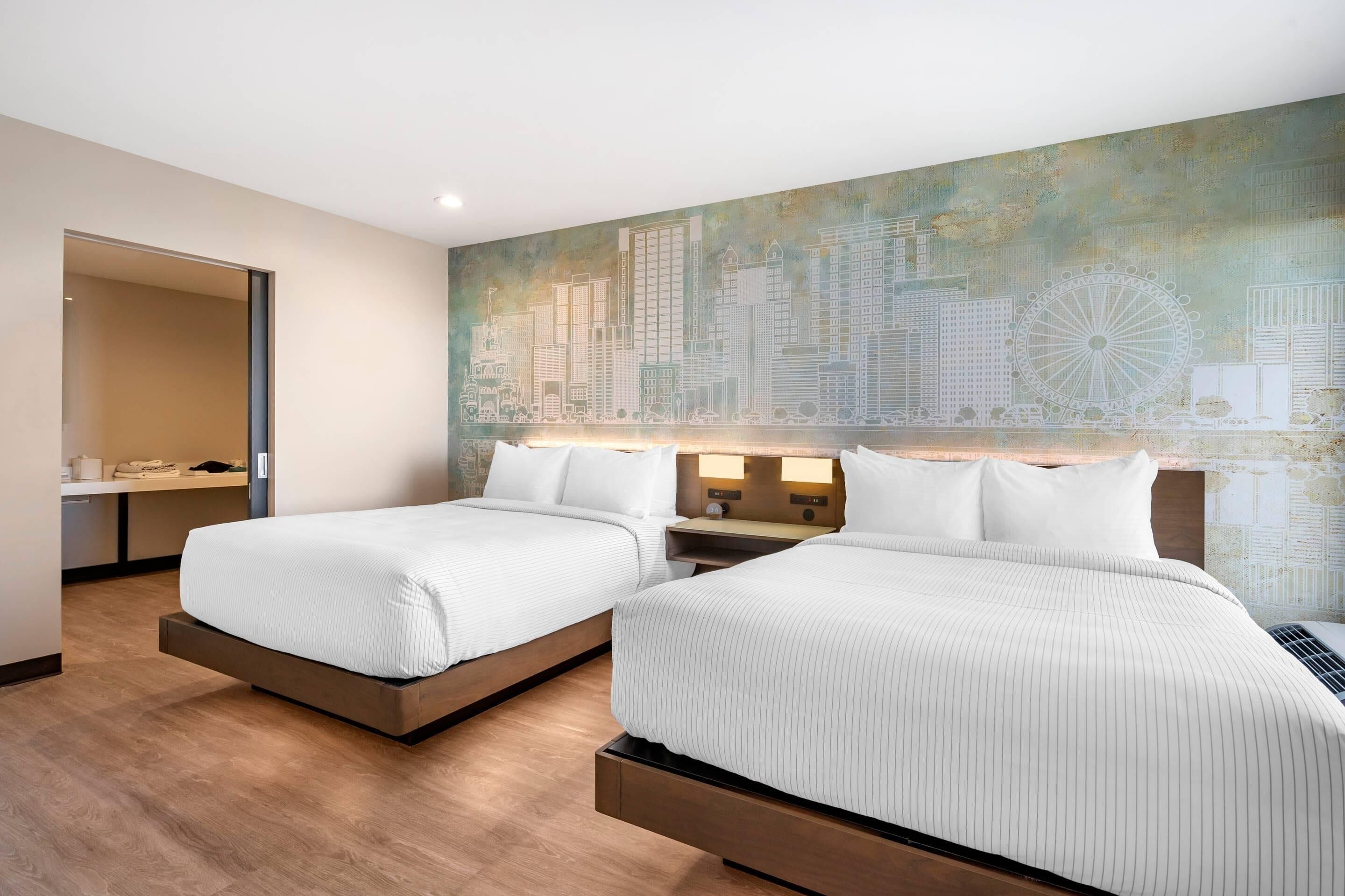 Una habitación de hotel con dos camas, un mural de paisaje urbano en la pared y suelo de madera.