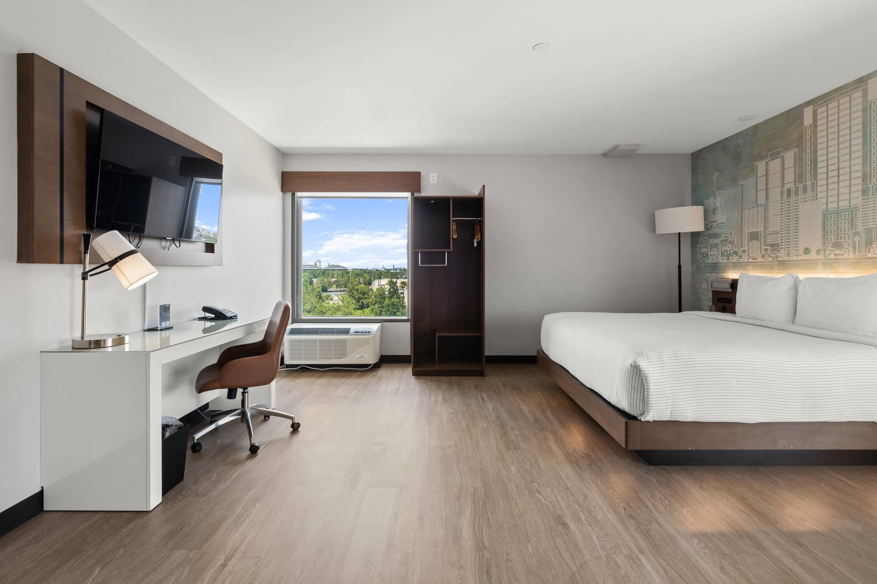 A modern hotel room with a large bed, a desk with a chair, a flat-screen TV, and a large window offering a city view.