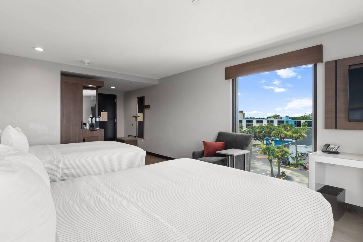 Una habitación de hotel moderna con dos camas, una pequeña zona de estar, un escritorio y un ventanal grande con vistas a la ciudad.
