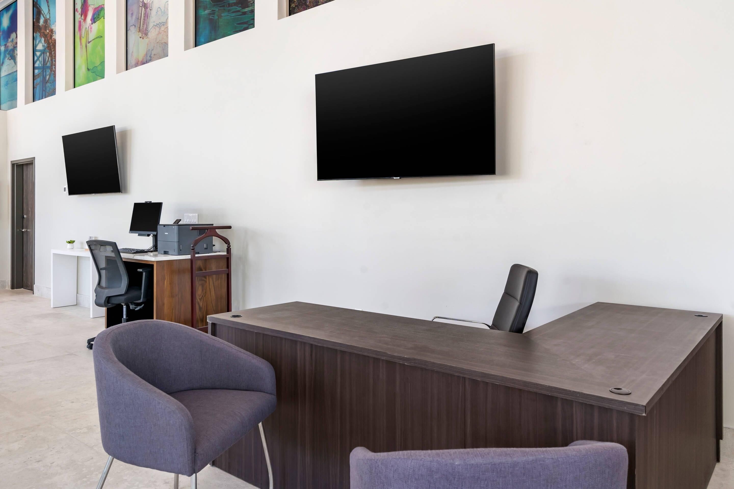 A modern office space with a reception desk, two chairs, a computer desk, and two flat-screen TVs mounted on the wall.