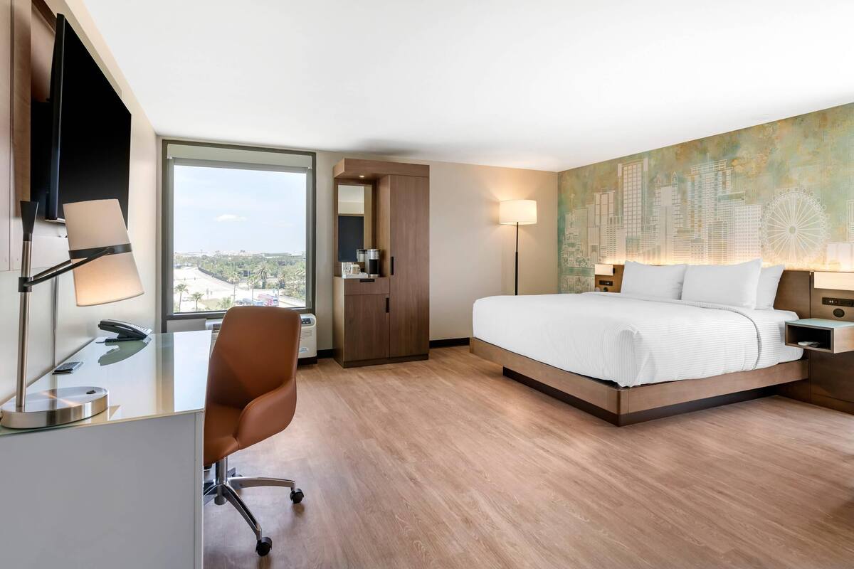 Una habitación de hotel moderna con una cama grande, un escritorio con una silla, una cafetera y vistas a la ciudad por la ventana.