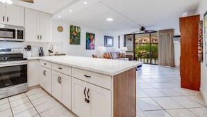 A modern kitchen with white cabinetry, a large island, and a dining area with a view of the outdoors. - Ground Floor Condo w/Kitchen - Kamaole Sands 3102 (Kihei)