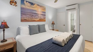 Ein Schlafzimmer mit einem Bett, zwei Nachttischlampen, einem Deckenventilator, einem Strandbild und einem durchgegangene Tür sichtbaren Badezimmer.