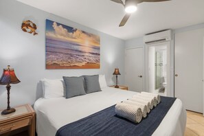 Ein Schlafzimmer mit einem Bett, zwei Nachttischlampen, einem Deckenventilator, einem Strandbild und einem durchgegangene Tür sichtbaren Badezimmer.