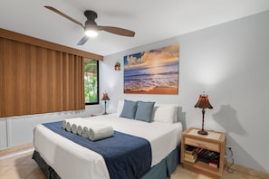Un dormitorio con una cama, un ventilador de techo, un cuadro de playa y una mesita de noche con libros.