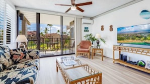 A spacious living room with a large sliding glass door leading to a balcony, a ceiling fan, a sofa with a floral pattern, a coffee table, and a TV on a wooden stand. - Kamaole Sands 1 BR Partial Ocean View #2-302 (KIHEI)