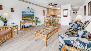 A spacious living area with a ceiling fan, wicker furniture, a television, and a kitchen in the background. - Kamaole Sands 1 BR Partial Ocean View #2-302 (KIHEI)