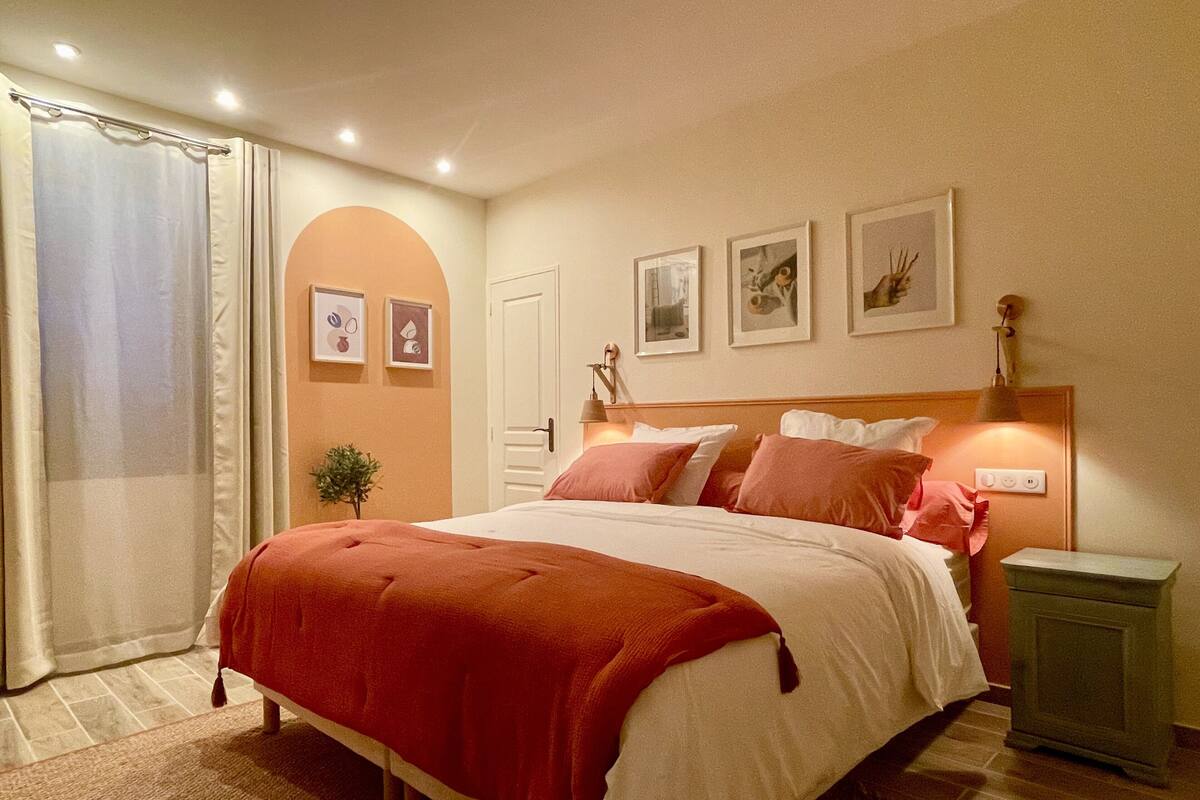 A bedroom with a bed, a nightstand, a lamp, a door, and framed pictures on the wall.
