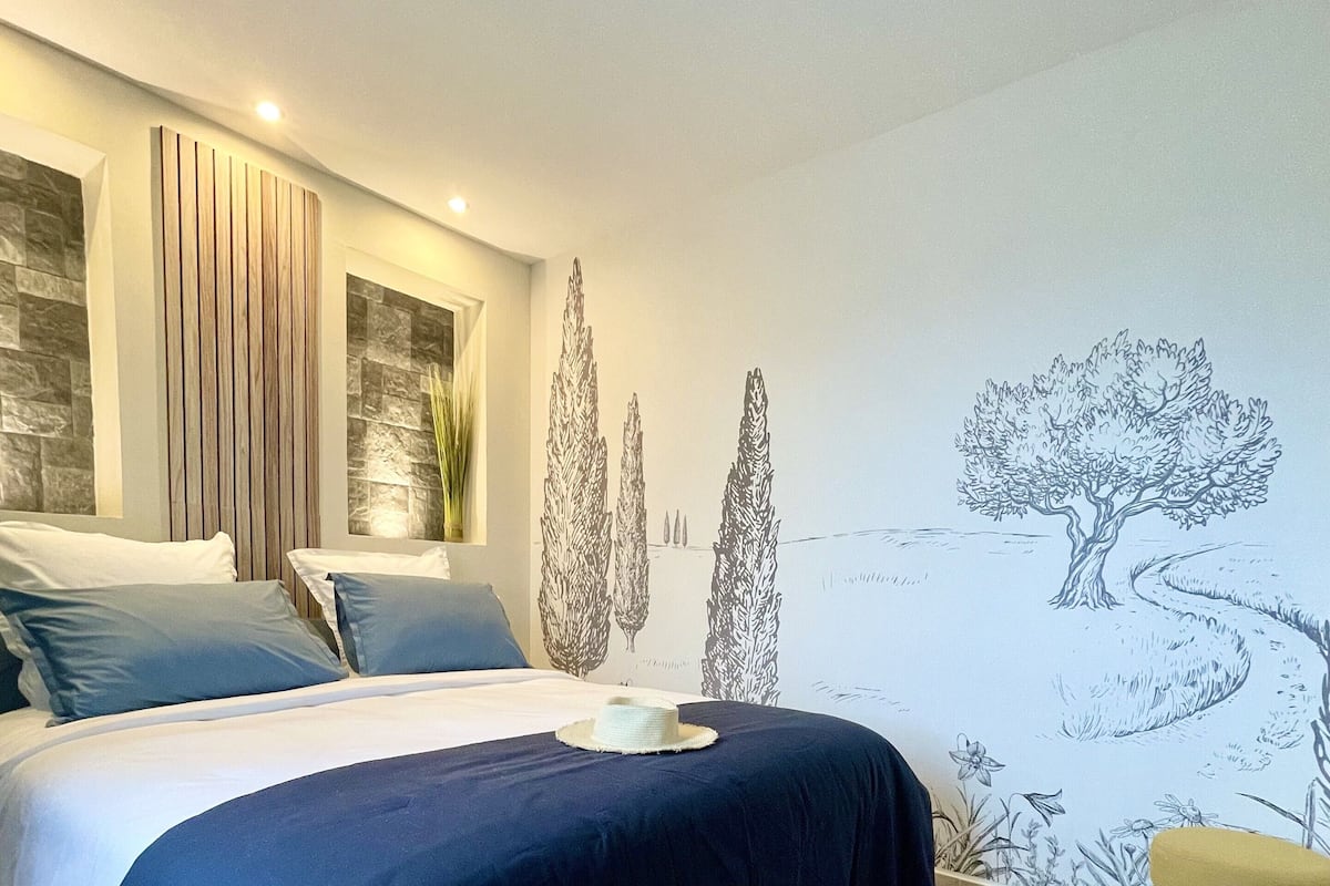 A modern bedroom with a bed, a nightstand, a wall mural, and a small stool.