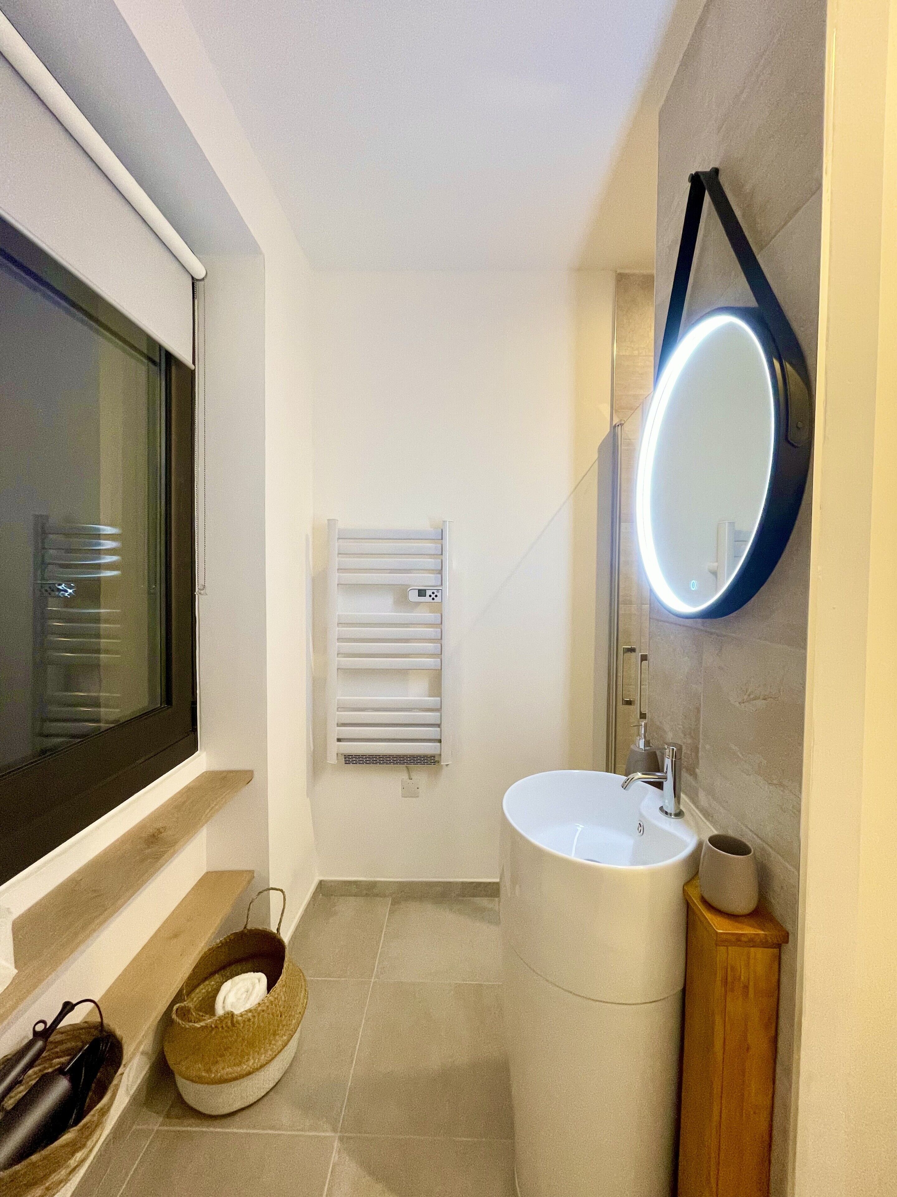 A modern bathroom with a large window, a built-in heater, a round mirror, and a sink.