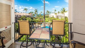 A balcony with two wicker chairs and a table, overlooking a resort area with palm trees and a clear sky.