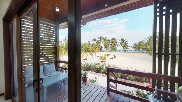A wooden deck with a bench and a table, overlooking a beach with palm trees and a clear sky.