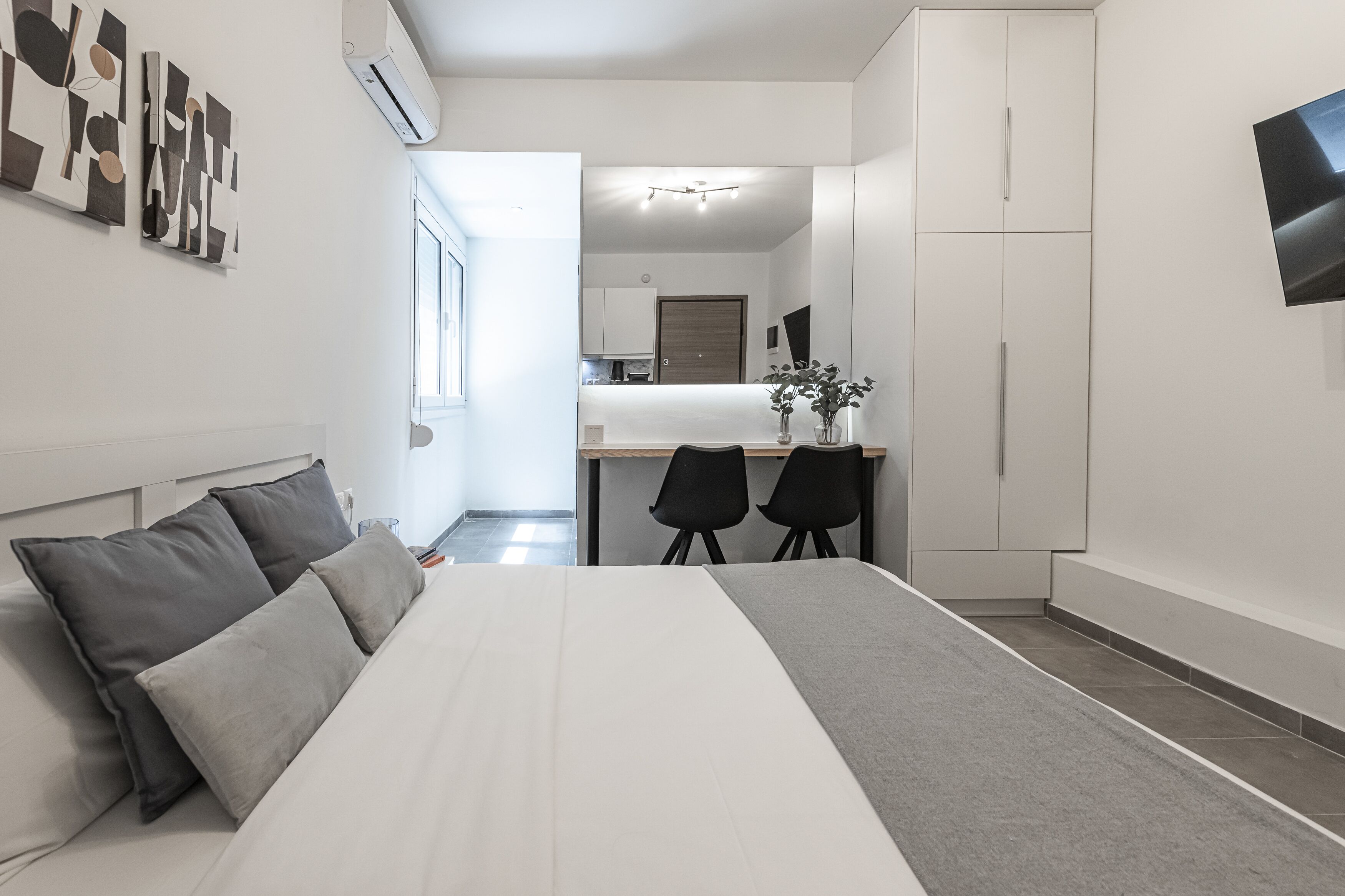 A modern bedroom with a bed, a desk, and chairs. There is a kitchen in the background.