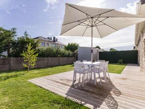 A patio with a table and chairs under a large umbrella, surrounded by a wooden deck and a grassy lawn.