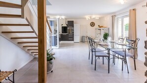 Una cocina moderna con armarios blancos, una zona de comedor con mesa de vidrio y sillas de metal, y una escalera de madera.