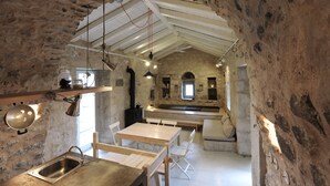 Un intérieur rustique en pierre, avec une table à manger en bois, des bancs et un coin cuisine doté d’un mur en pierre et d’une fenêtre.