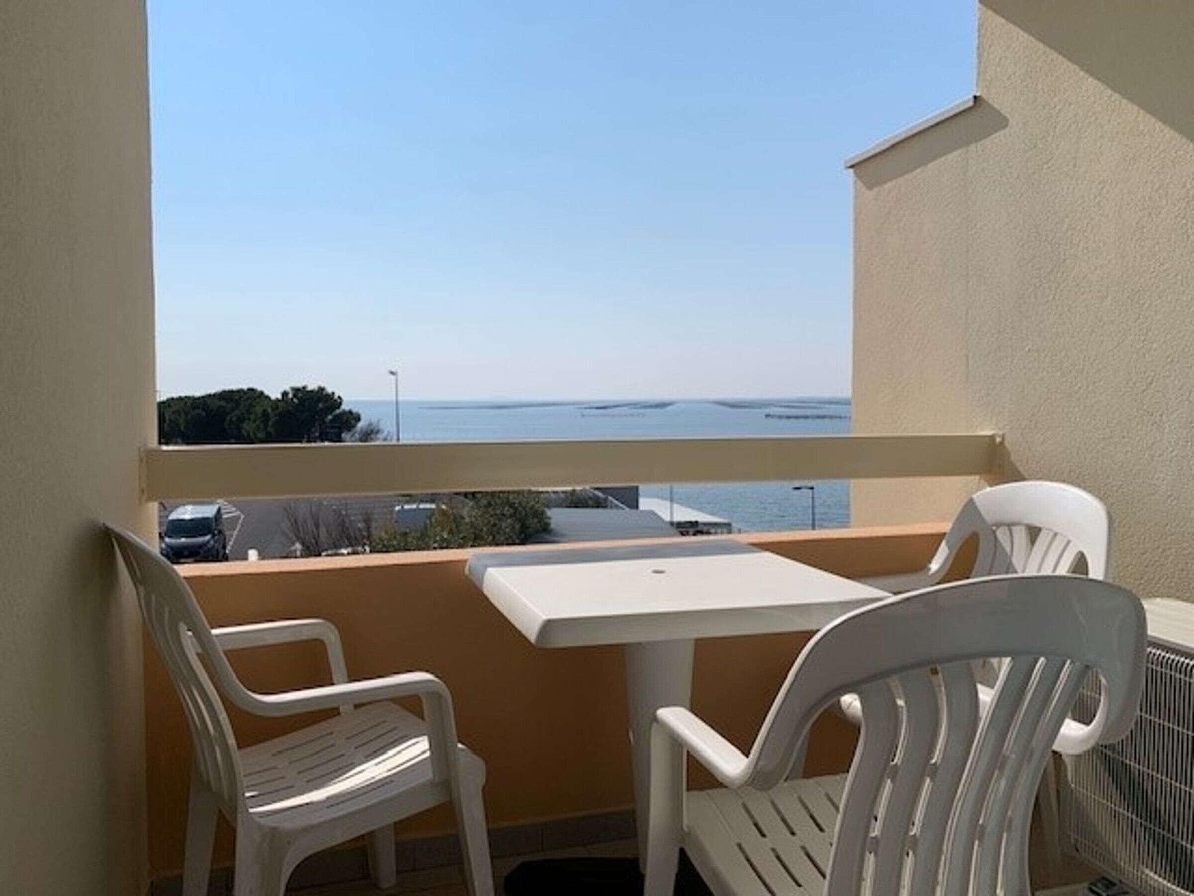 A balcony with a white table and two white chairs, overlooking a body of water and a parking area.
