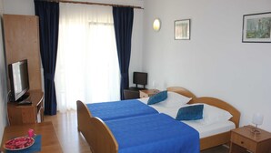 Quarto de hotel com duas camas, uma escrivaninha com televisão, uma cadeira, uma mesa de cabeceira e uma janela com cortinas.