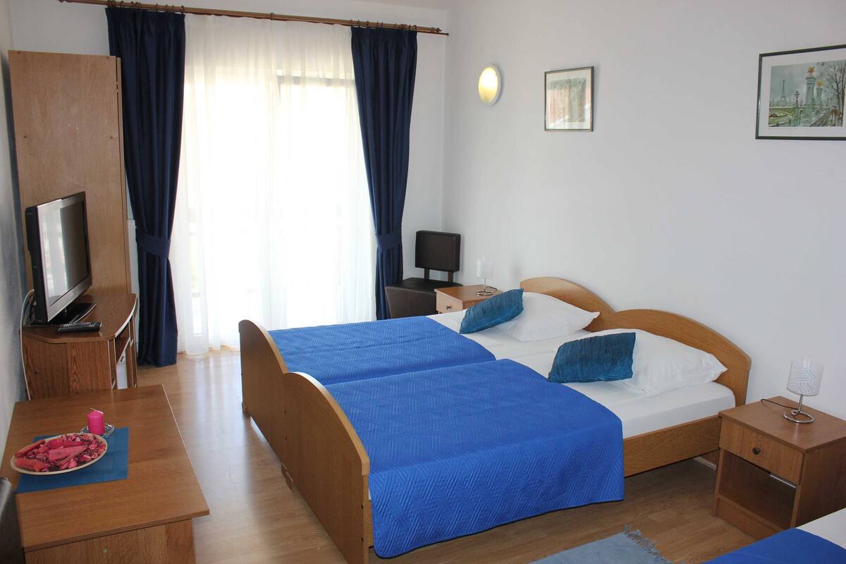 Ein Hotelzimmer mit zwei Betten, einem Schreibtisch mit Fernseher, einem Sessel, einem Nachttisch und einem Fenster mit Vorhängen.