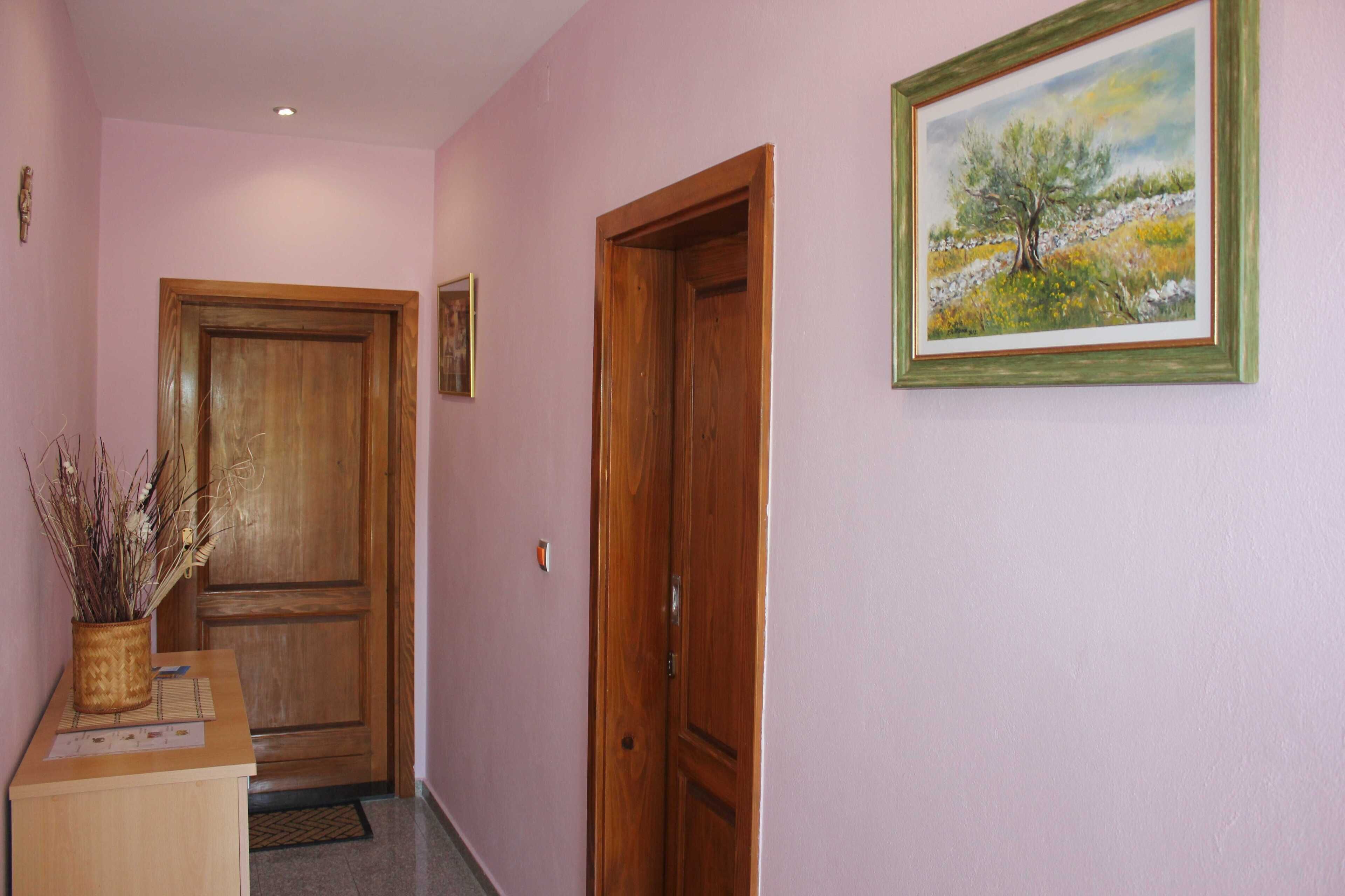 A hallway with a wooden door, a framed painting, and a desk with a vase of dried plants.