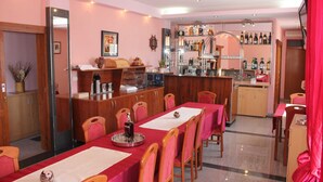 Uma sala de jantar com uma mesa comprida, cadeiras, um bar com estantes cheias de garrafas e uma cozinha ao fundo.