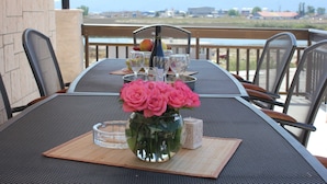 Uma mesa com um vaso de rosas cor-de-rosa, copos de vinho e uma garrafa, numa varanda com vista para montanhas e um corpo de água.