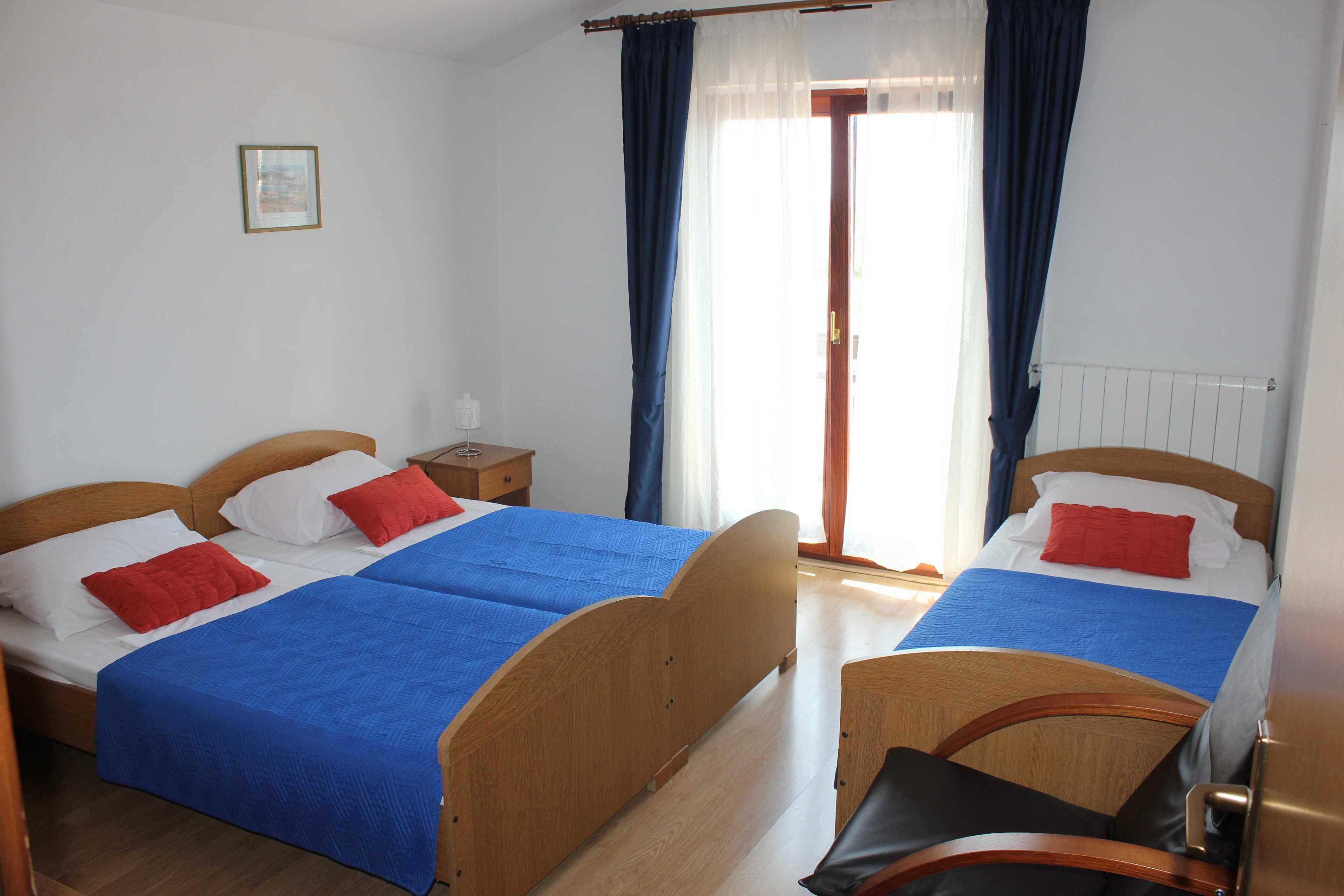 A double room with two beds, each with a blue bedspread and red pillows. There is a wooden headboard, a nightstand, and a window with curtains.