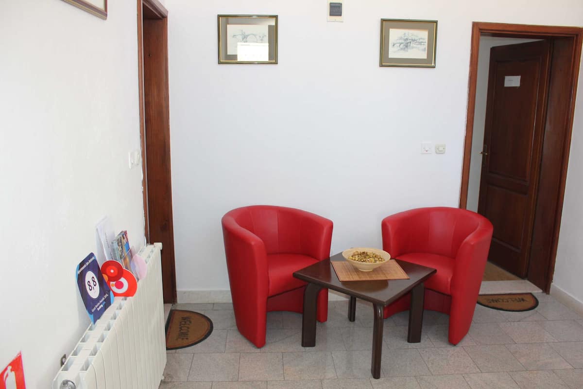 Ein Zimmer mit zwei roten Stühlen, einem kleinen Tisch und einem Heizkörper. An der Wand befinden sich gerahmte Bilder und eine Tür mit einem Schild.