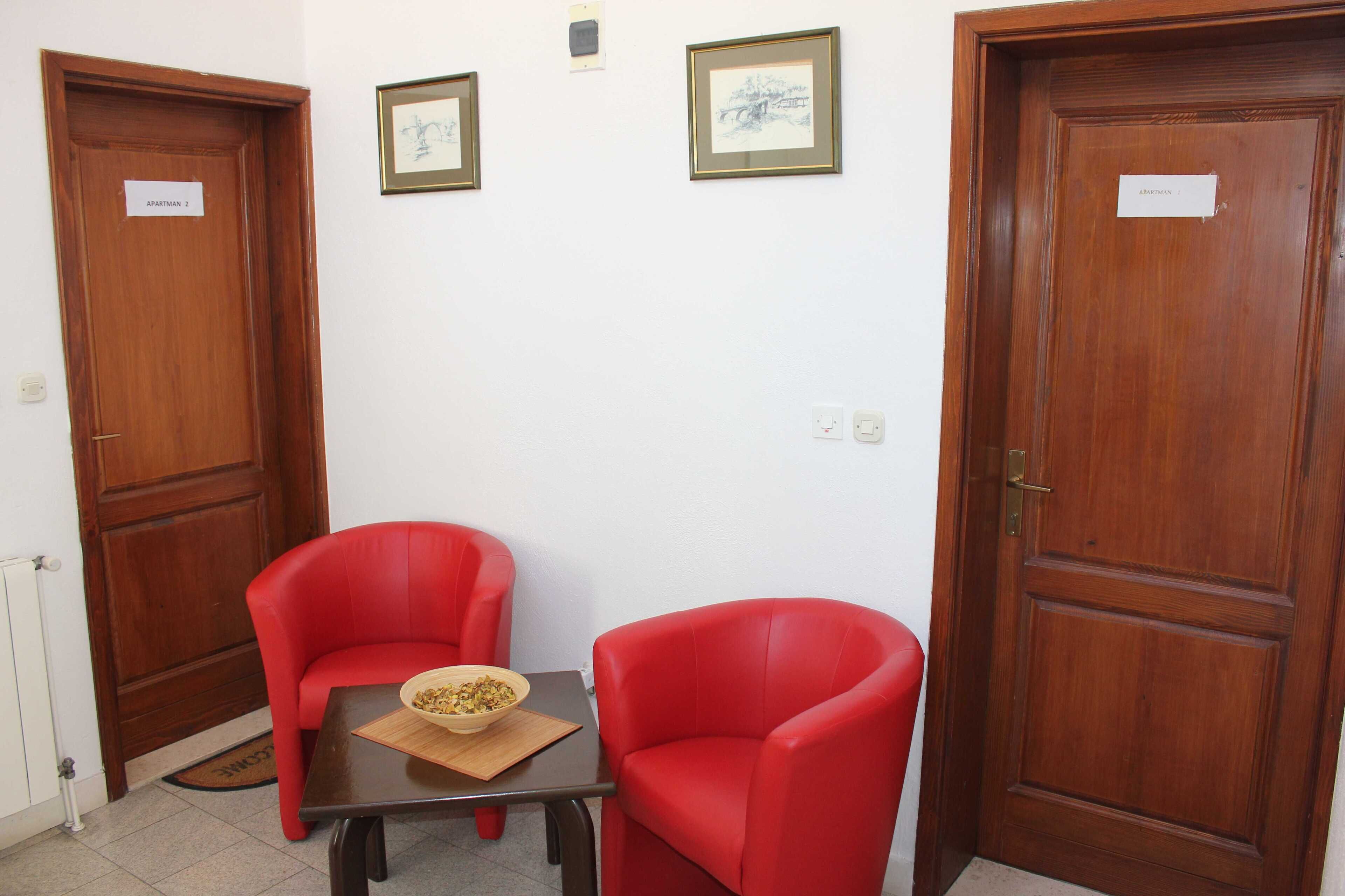 Una habitación con dos sillas rojas, una mesita con un bol, y dos puertas de madera con la leyenda 'Apartments 2'.
