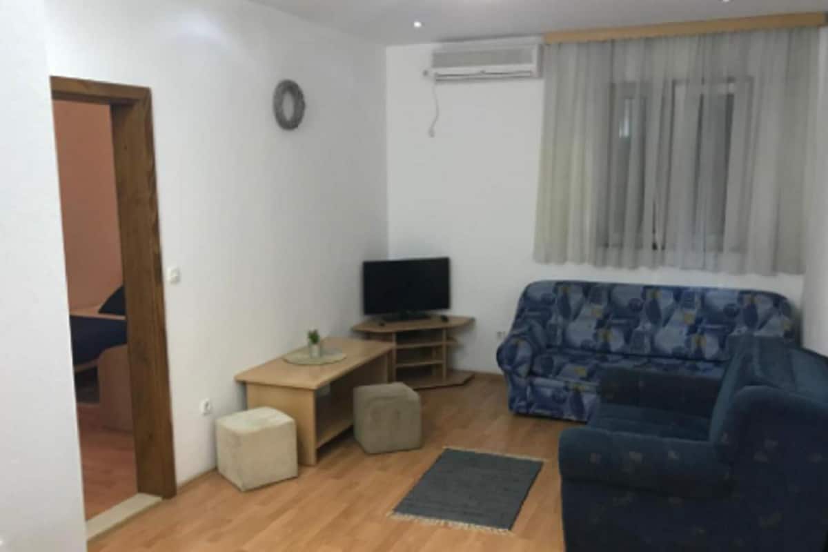 Ein Wohnzimmer mit Sofa, Fernseher auf einem Schrank, einem kleinen Tisch und einem Fenster mit Vorhängen.