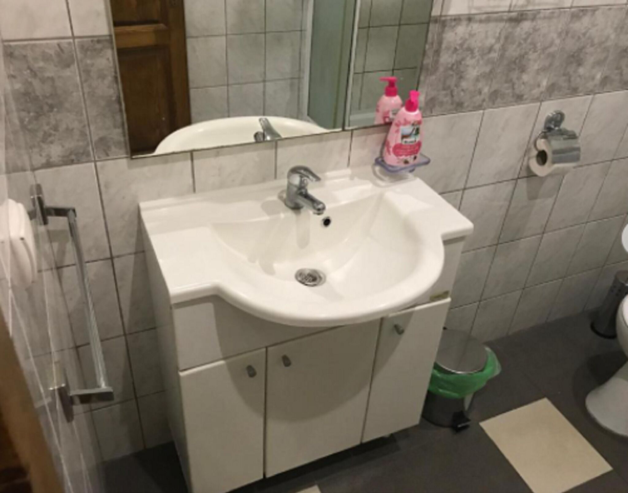 Un baño con lavamanos blanco, un espejo y un cubo de basura verde.