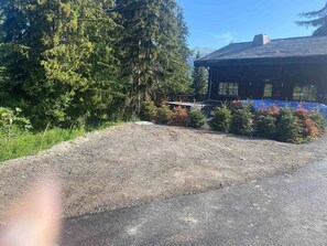 A gravel driveway leading to a building with a dark roof and windows, surrounded by evergreen trees and a blue fence.