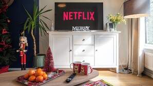 Ein Wohnzimmer mit einem Fernseher, der "NETFLIX" und "FRÖHLICHE WEIHNACHTEN" anzeigt, einer kleinen Weihnachtstanne, einer Nussknackerfigur, einer Schale Orangen und einer Fernbedienung.