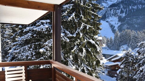 Un balcon en bois avec une chaise blanche, donnant sur un paysage montagneux enneigé, avec des arbres et des bâtiments.