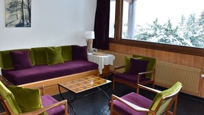 Une pièce chaleureuse avec un canapé vert et violet, deux chaises en bois, une petite table et une fenêtre donnant sur des arbres couverts de neige.