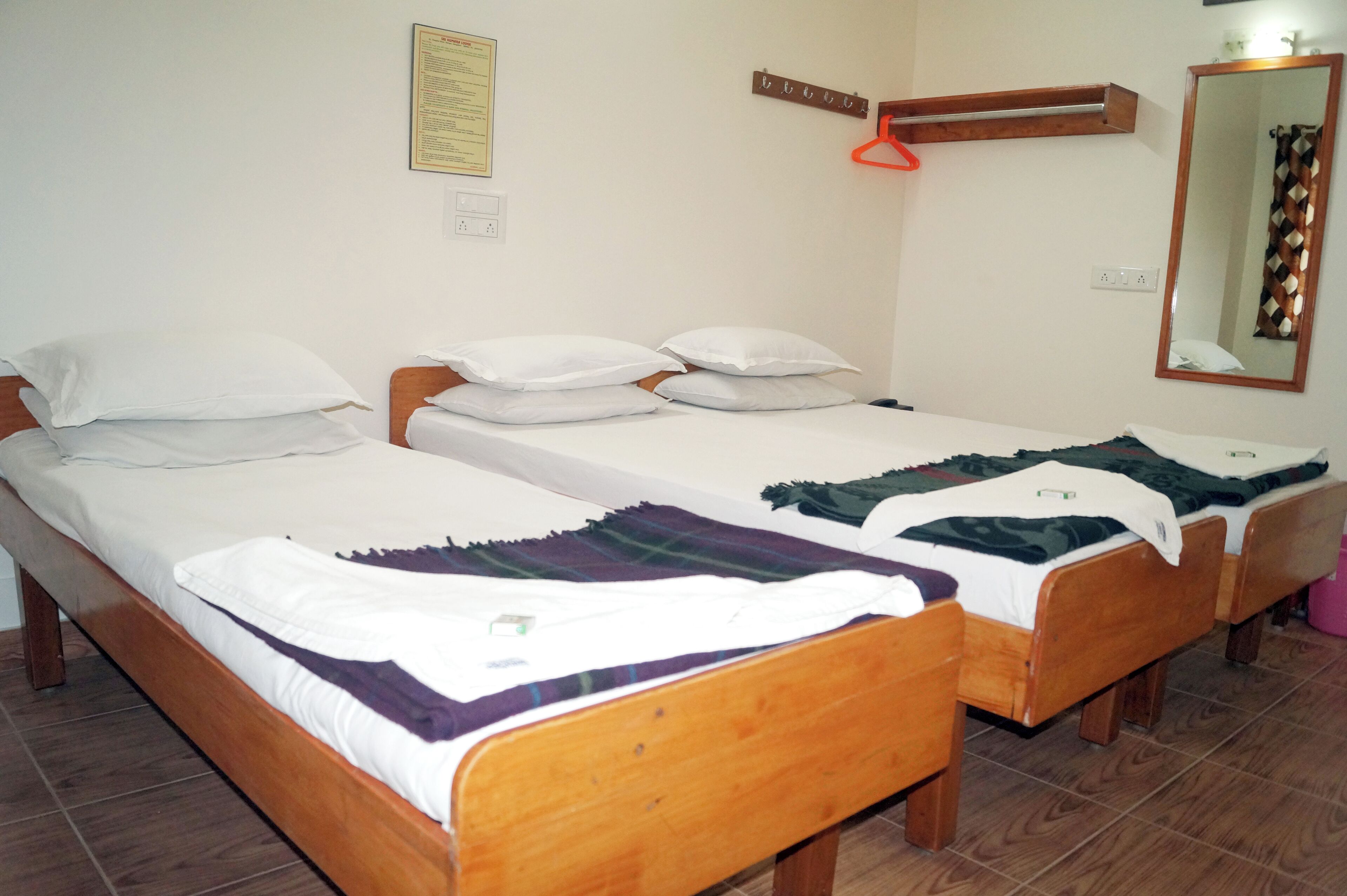 A room with two wooden beds, each with white bedding and a purple blanket. There is a wooden shelf with hooks on the wall, and a mirror with a decorative frame.