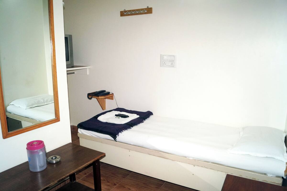 A small, simple room with a bed, a wooden nightstand, a mirror, and a wall-mounted shelf.