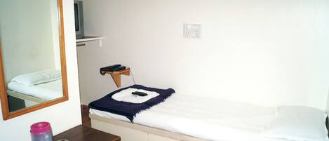 A small, simple room with a bed, a wooden nightstand, a mirror, and a wall-mounted shelf.