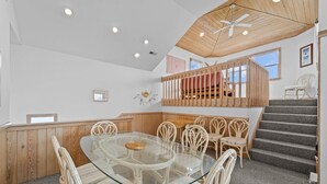 Un espace repas avec une table à plateau de verre entourée de chaises en osier, d’un banc en bois et d’un escalier menant à un niveau supérieur.