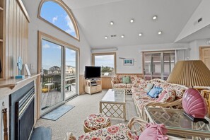 A spacious living room with a large window, a television, and a sofa with floral cushions. - 1609 - Barefoot Contessa - Walk to Beach | Hot Tub & Community Pool (Corolla)